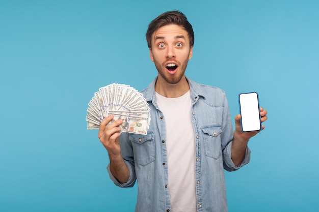 Portrait d'un homme surpris en chemise en denim de travailleur tenant des billets en dollars, un téléphone portable et regardant la caméra avec une expression naïve étonnée, choqué par la victoire des paris en ligne. tourné en studio intérieur isolé