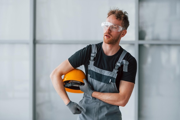 Portrait d'un homme en uniforme gris et avec un casque orange dans les mains qui se tient à l'intérieur dans un grand bureau moderne pendant la journée