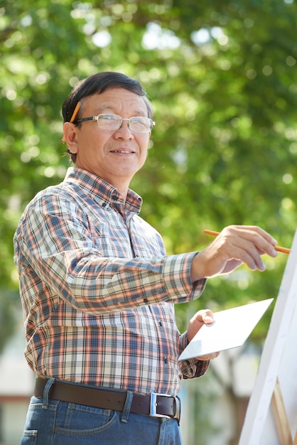 Portrait d'un homme vietnamien mature souriant à lunettes debout dans un parc et dessinant une photo avec des peintures