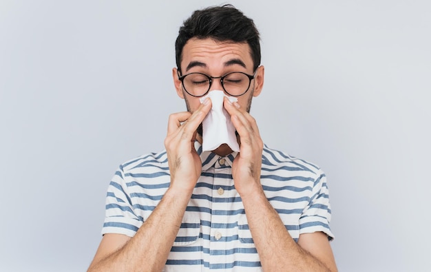 Portrait horizontal d'un bel homme malsain portant une chemise rayée et des lunettes se mouchant dans les tissus Les hommes ont le virus de la grippe ou une allergie sur fond blanc Concept de médecine et de personnes saines