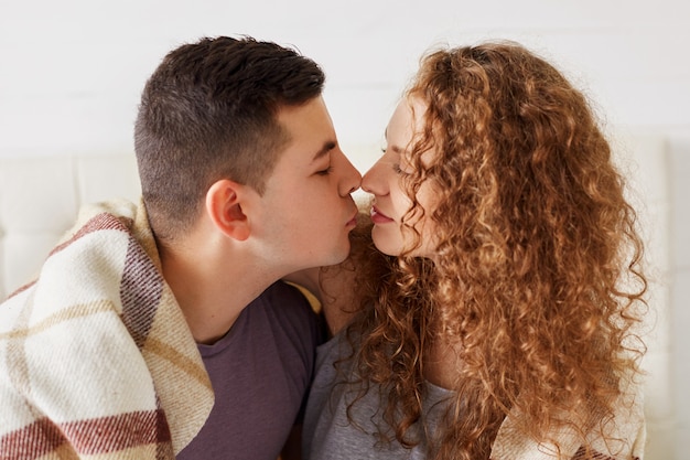 Portrait horizontal d'un couple de jeunes mariés affectueux toucher le nez et aller s'embrasser, profiter de l'atmosphère calme domestique, recouvert de plaid chaud