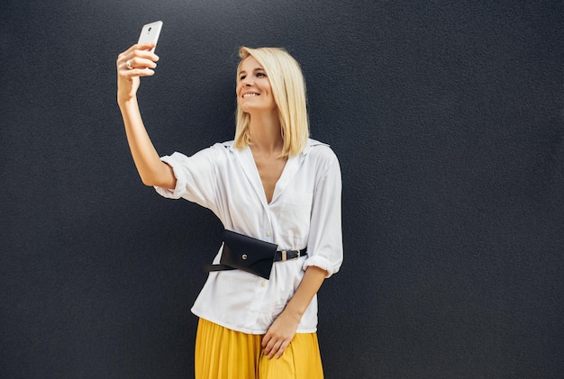 Portrait horizontal d'une femme heureuse debout à côté d'un mur gris tout en parlant d'autoportrait sur un téléphone intelligent Jolie femme porte une tenue décontractée et passe un appel en ligne sur un téléphone portable avec un ami Espace copie