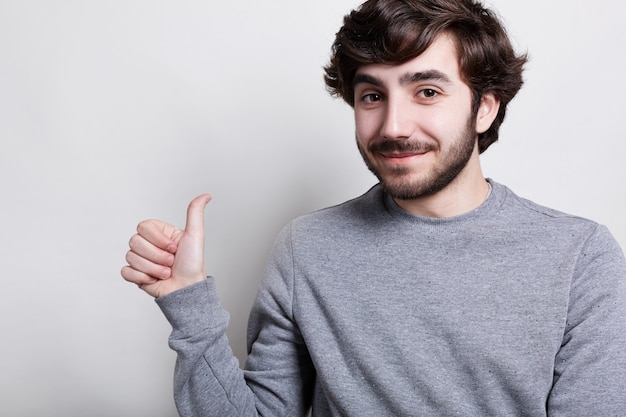 Un portrait horizontal de hipster barbu élégant avec une coiffure à la mode positive habillé en pull gris décontracté levant son pouce montrant un signe ok. Homme montrant sa satisfaction.