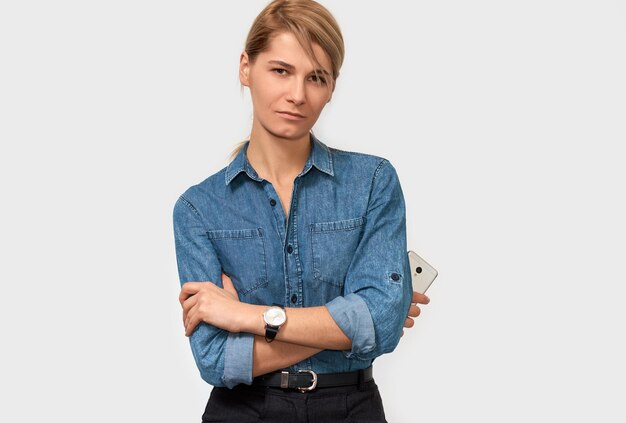 Portrait horizontal intérieur d'une pigiste blonde sérieuse aux cheveux blonds vêtue d'une chemise en jean bleu posant sur fond de studio Femme d'affaires avec téléphone portable posant sur un mur blanc