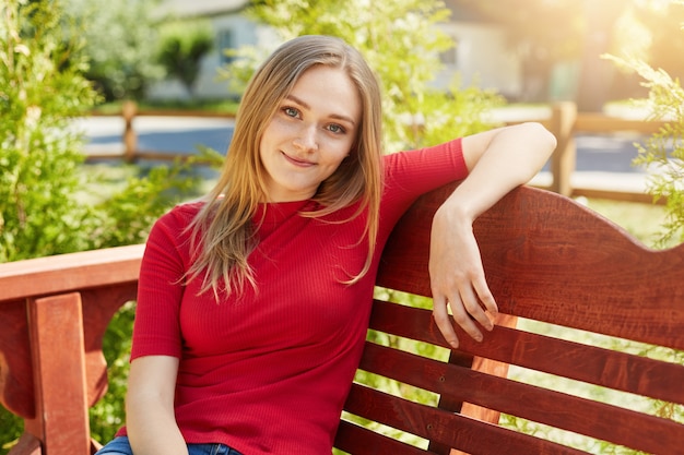 Portrait horizontal de jeune femme séduisante reposante avec de longs cheveux blonds ayant une apparence attrayante se sentant à l'aise tout en étant assis sur un banc en bois regardant agréablement dans l'appareil photo