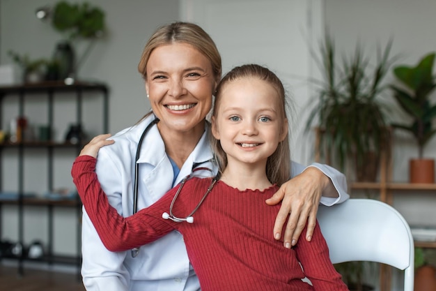 Photo portrait d'une infirmière amicale en blouse blanche embrassant avec un petit patient d'âge préscolaire heureux souriant ensemble à la caméra