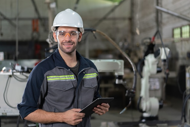 Portrait d'un ingénieur en automatisation masculin souriant tenir une tablette pour une machine de soudage à bras robotique d'inspection dans une usine industrielle