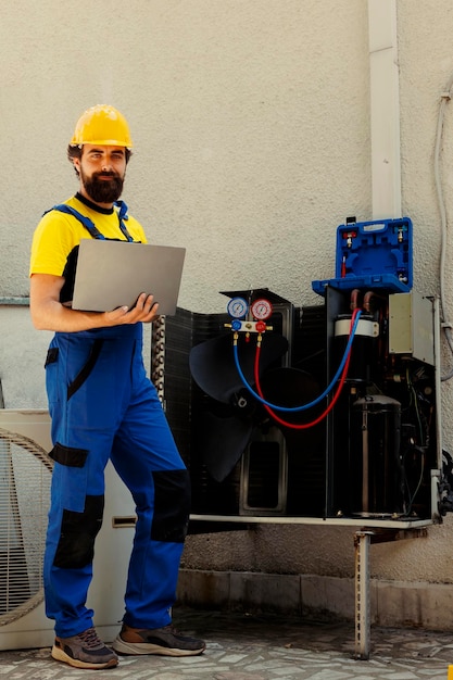 Portrait d'un ingénieur souriant et compétent travaillant sur un système CVC externe, tenant un ordinateur portable. Câbleur professionnel optimisant les performances du condenseur, garantissant qu'il fonctionne à sa capacité maximale