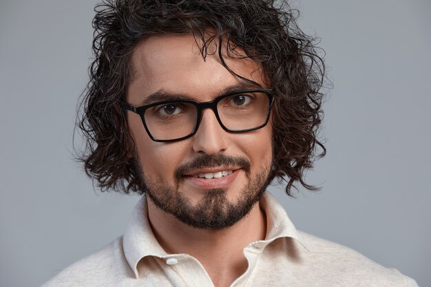 Portrait d'intérieur d'un beau mâle souriant aux taches de rousseur aux cheveux bouclés, porte des septtacles posant pour la publicité sociale, isolé sur un mur gris avec un espace de copie pour vos informations promotionnelles