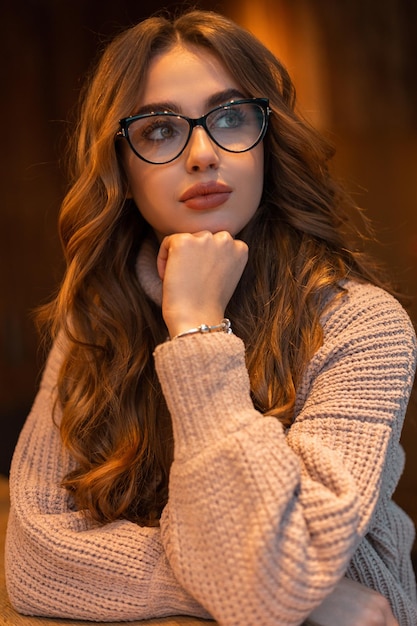 Portrait à l'intérieur d'une belle femme élégante avec des lunettes à la mode et une coiffure bouclée dans un pull vintage tricoté beige de mode se trouve dans un café