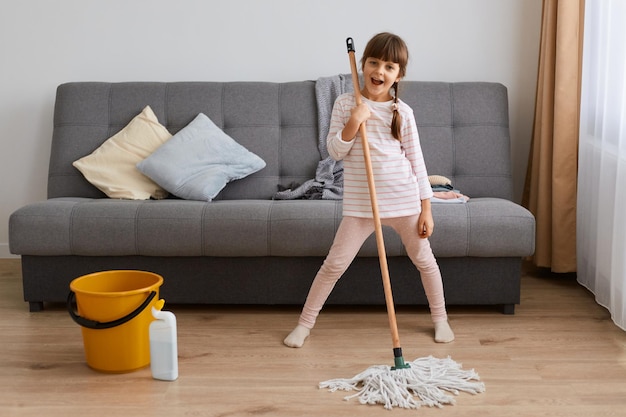 Portrait intérieur complet d'une petite fille avec des tresses vêtue d'une tenue décontractée debout près du sol du canapé et s'amusant à chanter en regardant la caméra en faisant seule les tâches ménagères