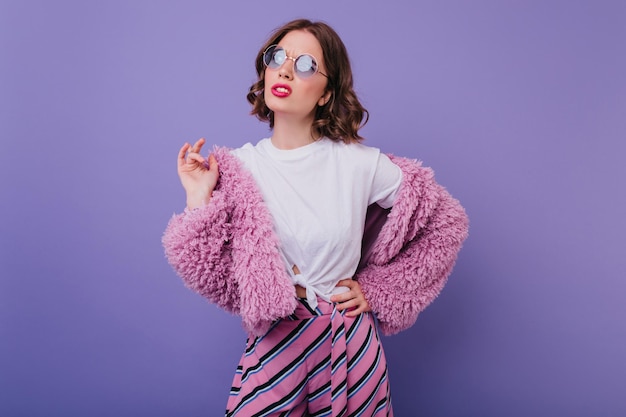 Portrait intérieur d'une fille bouclée bien habillée en lunettes de soleil isolées sur fond violet Photo en studio d'un merveilleux modèle féminin porte un pantalon rose et une veste moelleuse à la mode