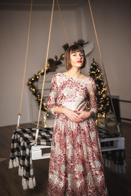 Portrait intérieur d'une fille confiante dans une longue robe posant près de l'arbre de Noël à la balançoire. Mise au point douce. Mise au point sélective.