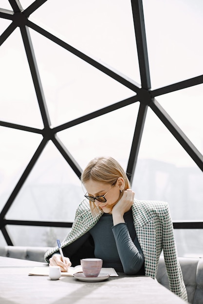 Portrait intérieur d'une jeune femme d'affaires assise dans un café et écrivant des documents. Fille blonde portant des lunettes de soleil et une veste