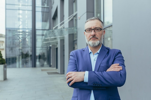 Portrait d'un investisseur senior aux cheveux gris sérieux avec les bras croisés à l'extérieur du bureau regardant