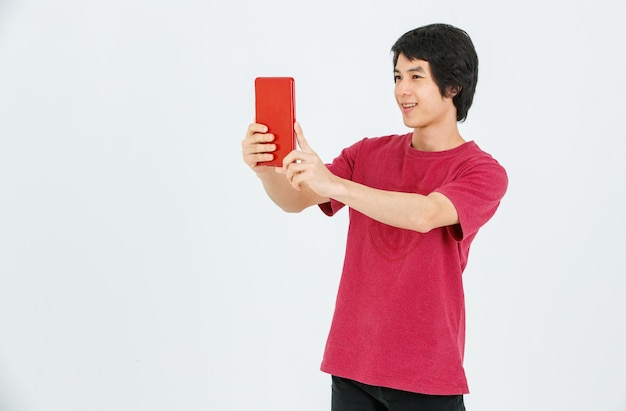 Portrait isolé coup de studio de découpe d'un modèle masculin asiatique jeune beau adolescent en tenue de style de rue debout tenant une tablette dans les mains souriant prenant une photo de selfie sur fond blanc