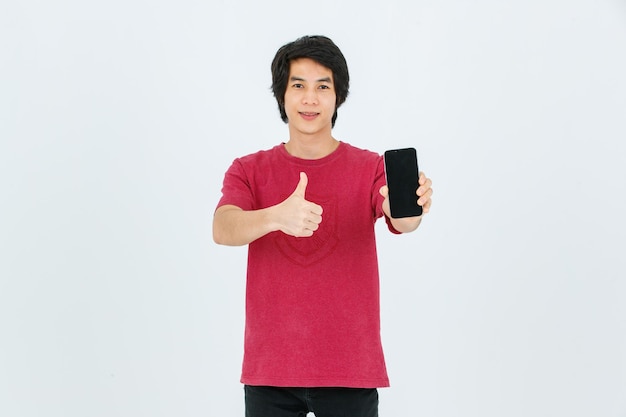 Portrait isolé découpe studio tourné asiatique jeune beau modèle masculin adolescent en tenue de style de rue debout souriant tenant un doigt pointé présentant un smartphone à écran blanc noir sur fond blanc
