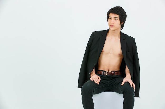Portrait isolé studio tourné asiatique jeune beau confiant mince mince torse nu sain athlétique adolescent mode modèle masculin en costume blazer jeans et baskets assis souriant posant sur fond blanc