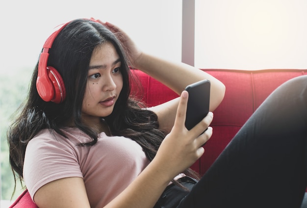 Portrait d'un jeune adolescent souriant et mignon dans des vêtements décontractés roses qui aiment écouter de la musique avec un casque et un smartphone. Jeune fille junior allongée sur le canapé rouge et jouant au téléphone portable