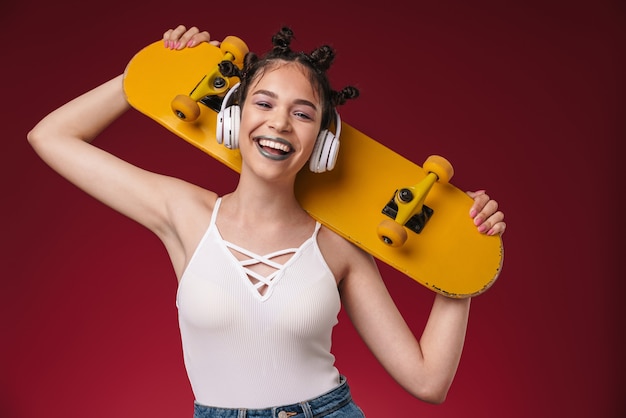 Portrait d'une jeune adolescente punk souriante écoutant de la musique avec des écouteurs isolés sur un mur rouge bordeaux tenant une planche à roulettes