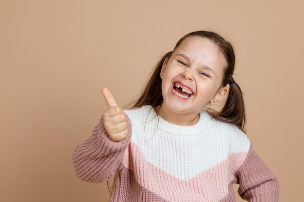 Portrait de jeune adorable fille riante joyeuse avec de longs cheveux noirs en pull rose blanc debout montrant les pouces vers le haut signe geste d'approbation sur fond marron