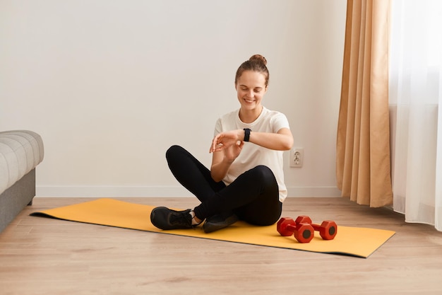 Portrait de jeune adulte belle femme caucasienne vérifiant le tracker de fitness numérique pendant l'auto-entraînement à la maison et souriant assis sur le sol sur un tapis de yoga
