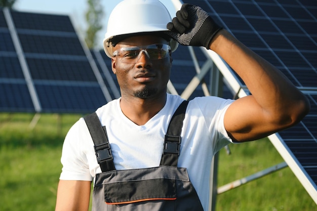 Portrait d'un jeune artisan afro-américain en casque de protection Homme en uniforme et avec des outils debout parmi les panneaux solaires