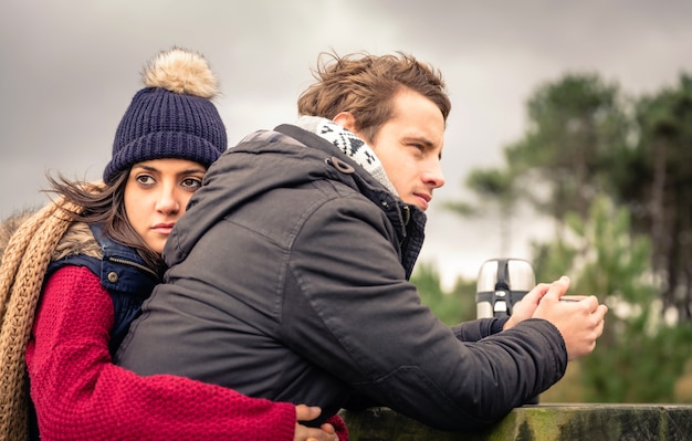 Portrait d'un jeune beau couple embrassant et buvant une boisson chaude par une journée froide avec un ciel sombre et nuageux en arrière-plan