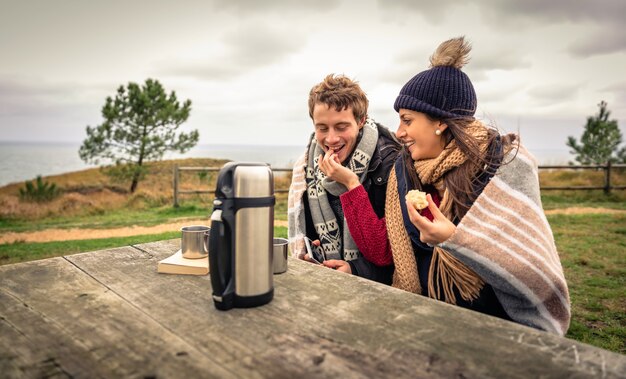 Portrait de jeune beau couple sous couverture mangeant des muffins dans une journée froide avec la mer et un ciel nuageux sombre sur l'arrière-plan