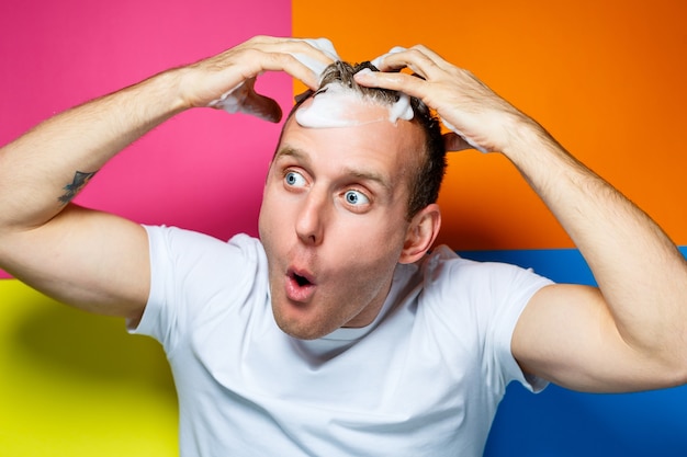 Photo portrait d'un jeune, beau et joyeux gars sur fond multicolore, vêtu d'un t-shirt blanc, crée une coiffure à la mode, des émotions stupides et positives