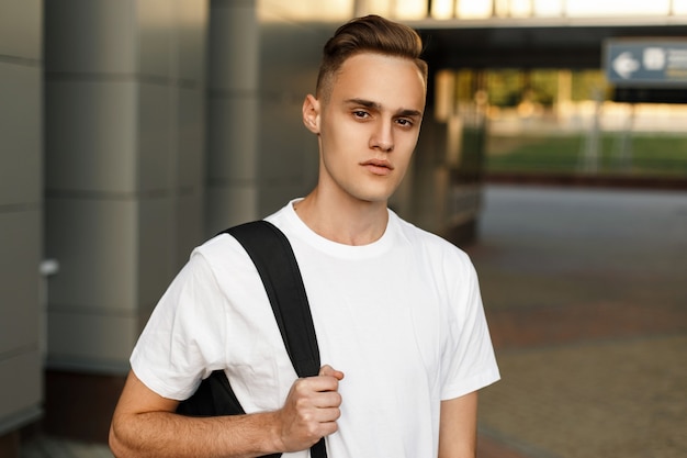 Portrait d'un jeune beau mec branché dans une chemise blanche avec un sac à dos près du bâtiment moderne.