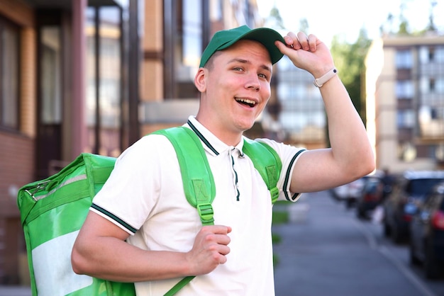 Portrait de jeune beau mec de messagerie heureux sourire livreur de nourriture avec une boîte thermo verte pour la livraison de nourriture à l'extérieur dans la cour sur fond de bâtiments à la journée d'été en casquette et uniforme