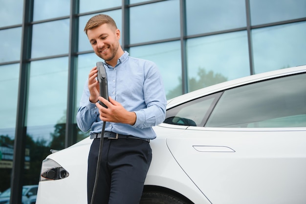 Portrait de jeune bel homme barbu en tenue décontractée debout à la station de charge et tenant une prise du chargeur pour une voiture électrique Concept de voiture électrique Eco