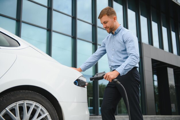 Portrait de jeune bel homme barbu en tenue décontractée debout à la station de charge et tenant une prise du chargeur pour une voiture électrique Concept de voiture électrique Eco