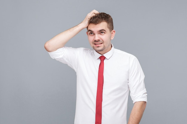 Portrait de jeune bel homme confus réfléchi en chemise blanche et cravate debout se grattant la tête et pensant. intérieur isolé sur fond gris.