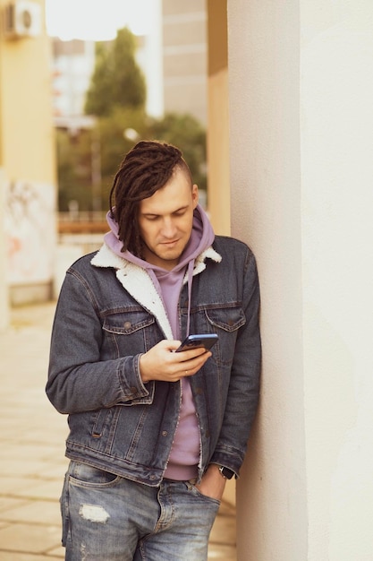 Portrait de jeune bel homme avec des dreadlocks naviguant sur un téléphone portable debout appuyé contre un mur dans la rue