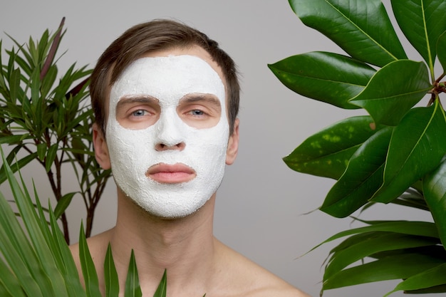 Portrait de jeune bel homme avec un masque cosmétique blanc sur son visage sur fond de plantes vertes. Soins du visage pour hommes