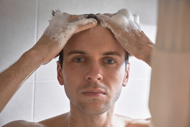 Portrait de jeune bel homme se lave avec du gel douche, mousse la tête avec du shampoing dans la salle de bain à la maison en gros plan