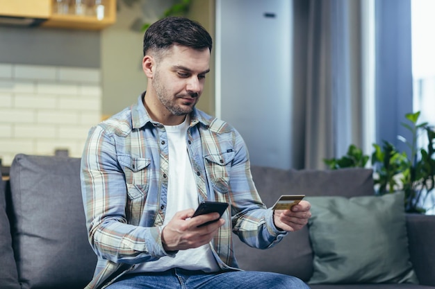 Portrait d'un jeune bel homme Tient un téléphone et une carte de crédit commande des marchandises fait des achats en ligne Assis à la maison sur le canapé regarde la chambre sourit