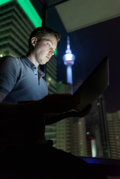 Portrait de jeune bel homme utilisant un ordinateur portable contre la fenêtre en verre avec vue sur la ville pendant la nuit