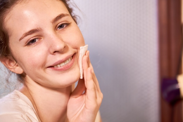 Portrait de jeune belle adolescente essuyant le visage propre avec du toner matifiant ou de l'eau micellaire