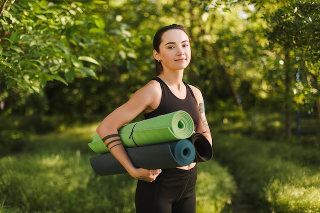 Portrait de jeune belle dame en haut sportif noir et leggings debout avec des tapis de yoga dans les mains et regardant rêveusement à huis clos dans le parc