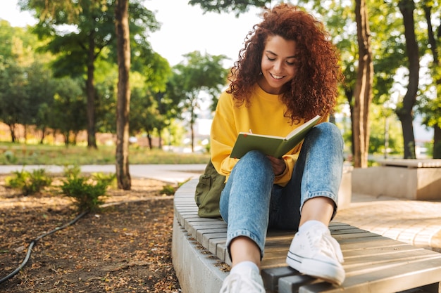 Portrait d'une jeune belle étudiante bouclée heureuse assise à l'extérieur dans un parc naturel, écrivant des notes dans un cahier.