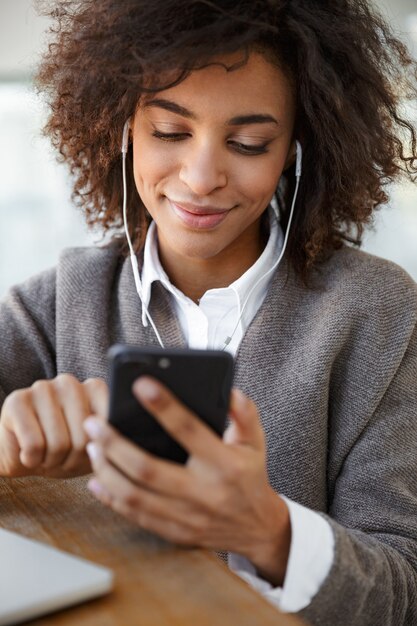 Photo portrait de jeune belle femme afro-américaine à l'aide d'un ordinateur portable et d'un téléphone portable assis près de la fenêtre du café