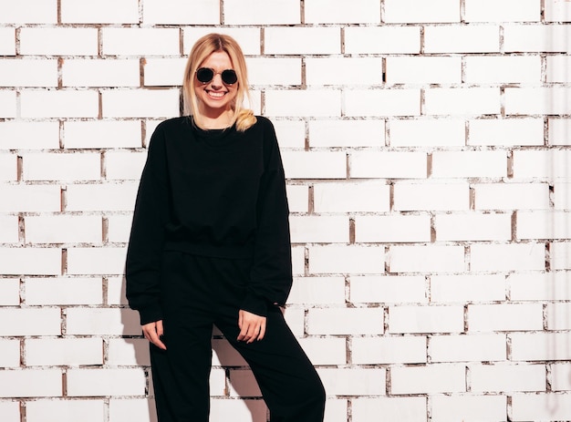 Portrait de jeune belle femme blonde souriante dans des vêtements surdimensionnés noirs d'été à la mode Sexy femme insouciante posant près d'un mur de briques blanches en studio Modèle positif s'amusant à l'intérieur