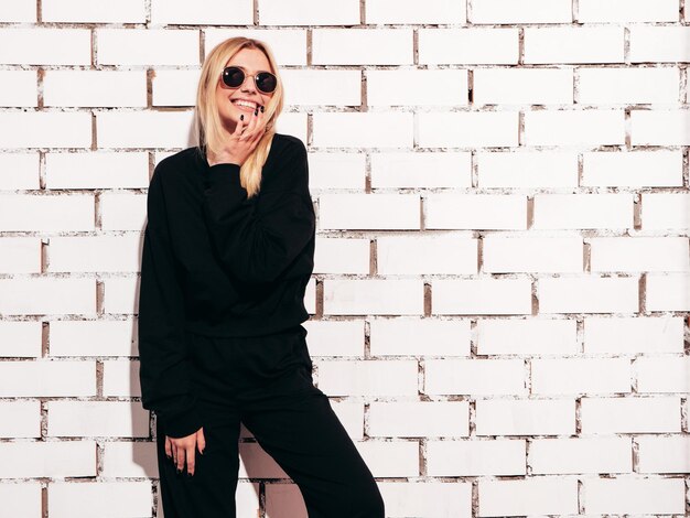 Portrait de jeune belle femme blonde souriante dans des vêtements surdimensionnés noirs d'été à la mode Sexy femme insouciante posant près d'un mur de briques blanches en studio Modèle positif s'amusant à l'intérieur