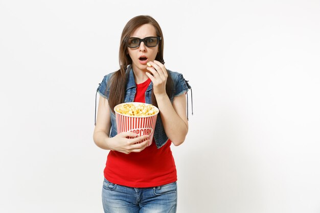 Portrait de jeune belle femme brune choquée dans des lunettes 3d, vêtements décontractés regardant un film, tenant et mangeant du pop-corn dans un seau isolé sur fond blanc. Émotions dans le concept de cinéma.