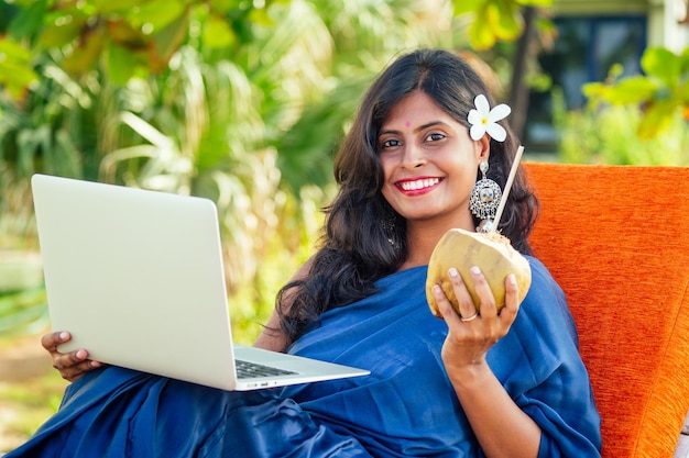 Portrait de jeune belle femme indienne avec plumeria de fleurs blanches en entendant boire de l'eau de noix de coco et travaillant par ordinateur portable dans la mer de Goa en Inde Elle utilise un tube recyclable en métal écologique dans une station thermale