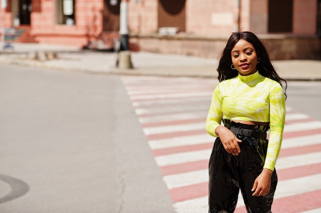 Portrait de jeune belle femme mannequin afro-américaine marchant sur un passage pour piétons, porter un haut vert et un pantalon noir brillant et mouillé.