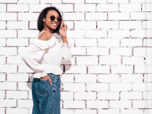 Portrait de jeune belle femme noire Modèle souriant habillé en jeans d'été Sexy femme insouciante posant près d'un mur de briques blanches en studio Bronzé et joyeux